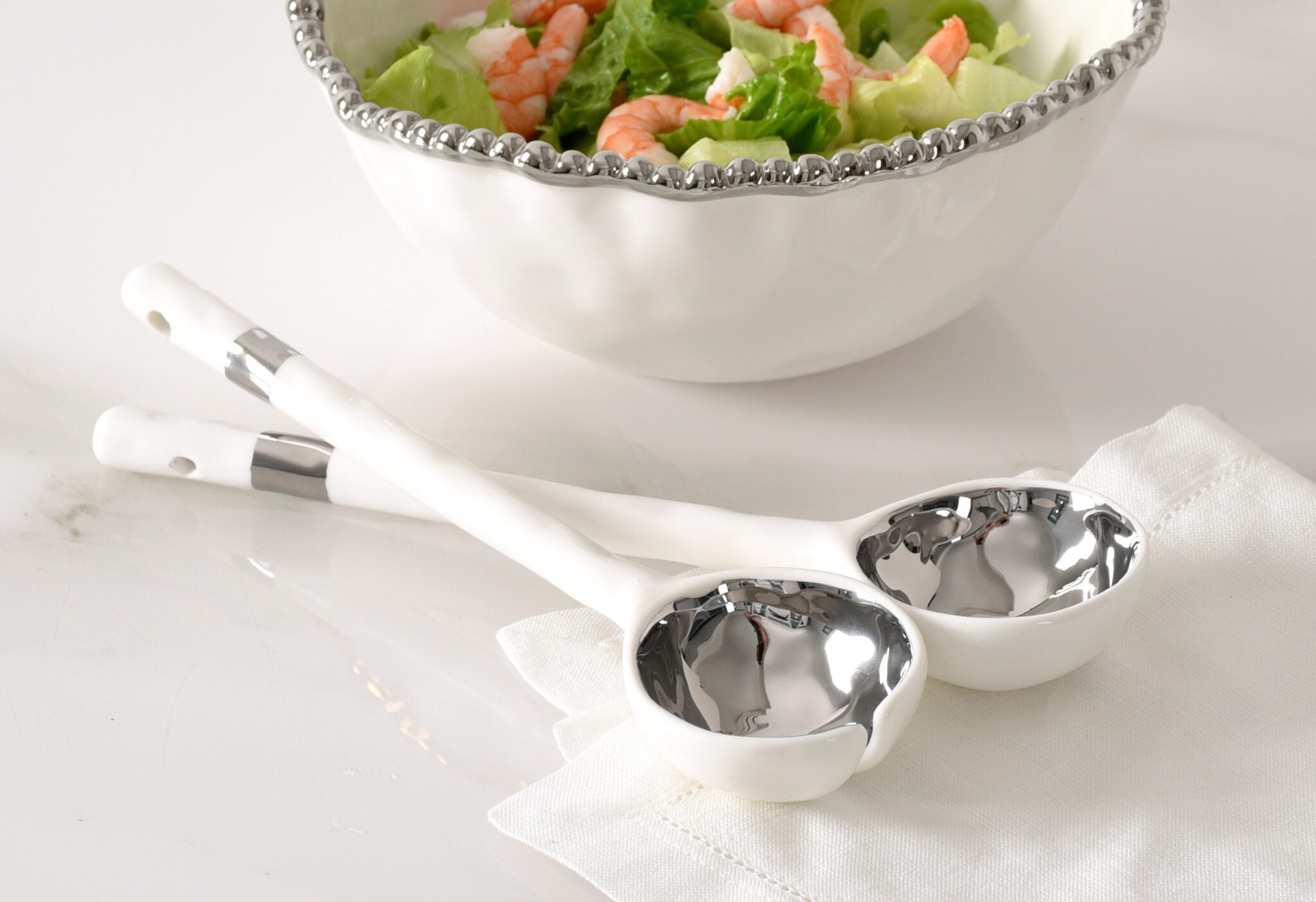 Two metal serving spoons on a white napkin with a bowl of salad in the background.