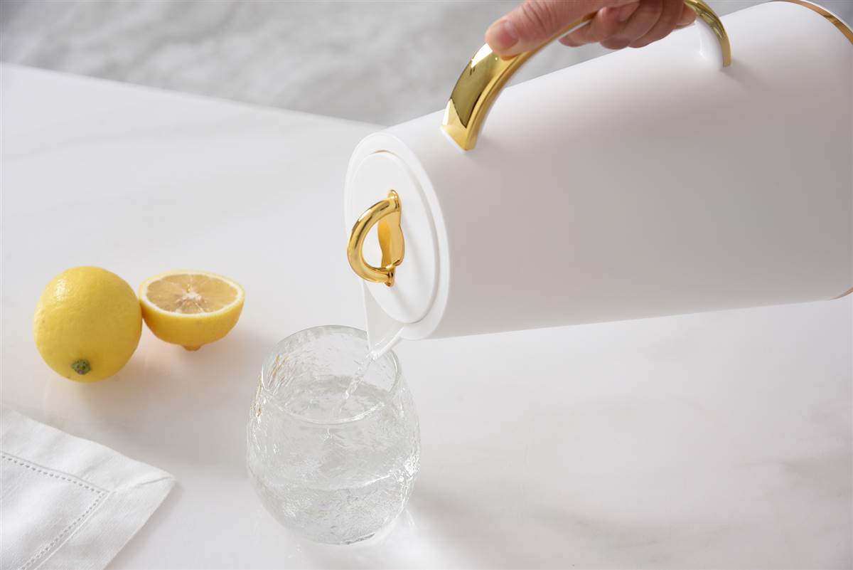 White carafe with a gold handle pouring water inot a glass cup next to lemons. 