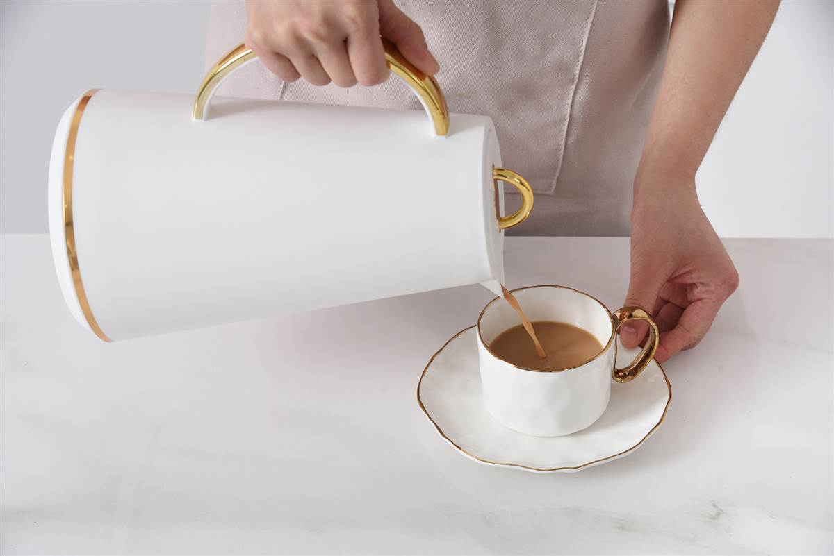 Person pouring coffee from a white pitcher with gold accents into a matching cup and saucer on a light surface.
