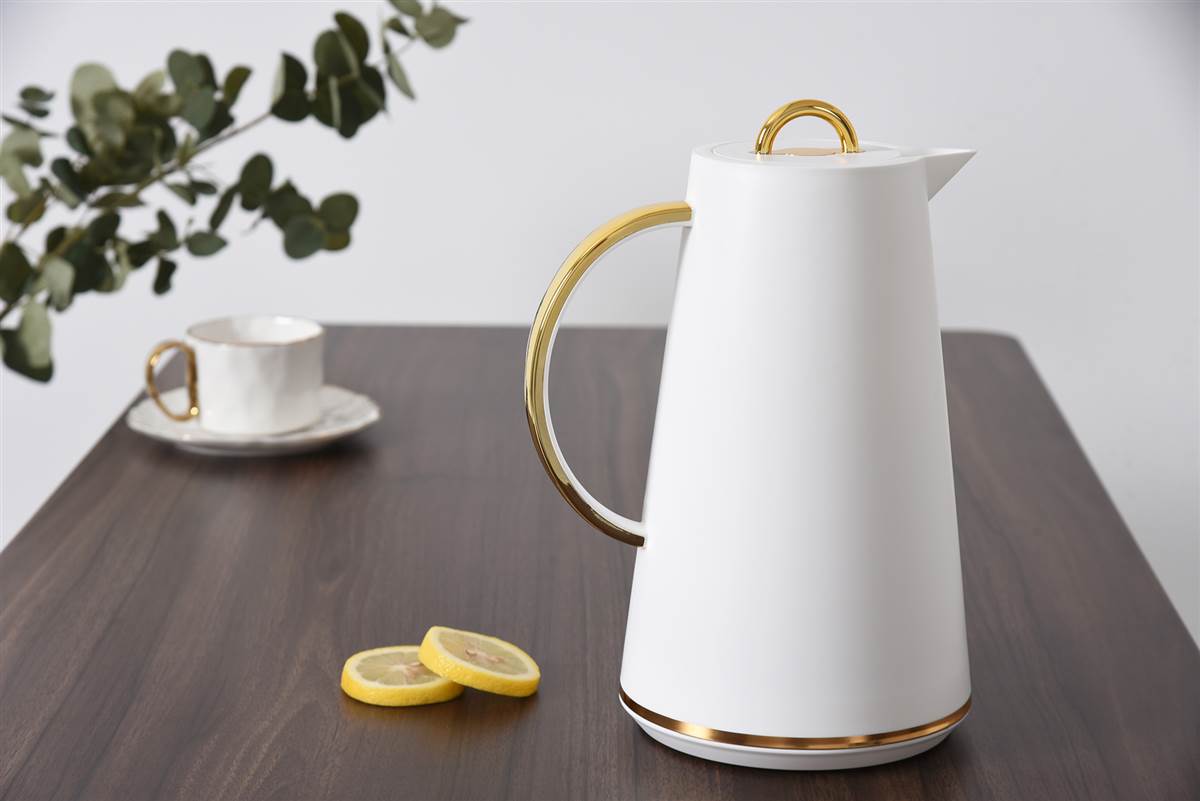 White pitcher with gold accents on a wooden table with lemon slices and a cup.
