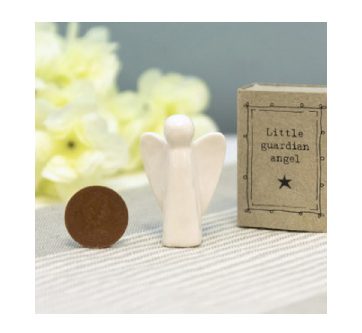 Small wooden angel figurine next to a block labeled 'Little guardian angel' on a textured surface with blurred flowers in the background.