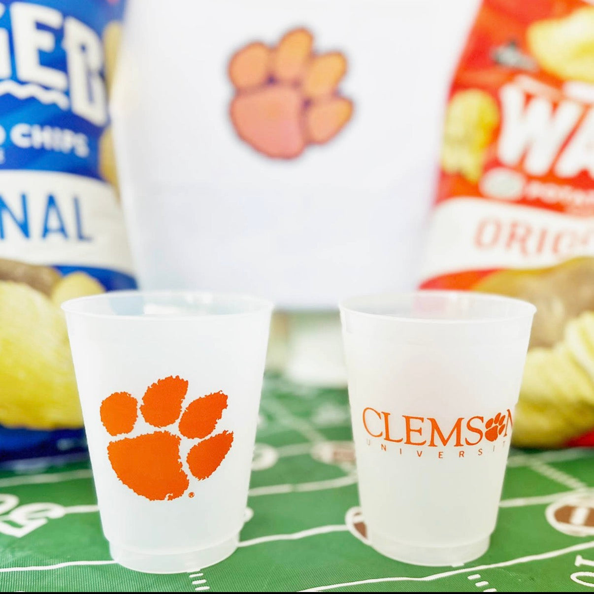 Two frosted shot glasses with a paw print design on a sports-themed tablecloth, surrounded by snack bags.