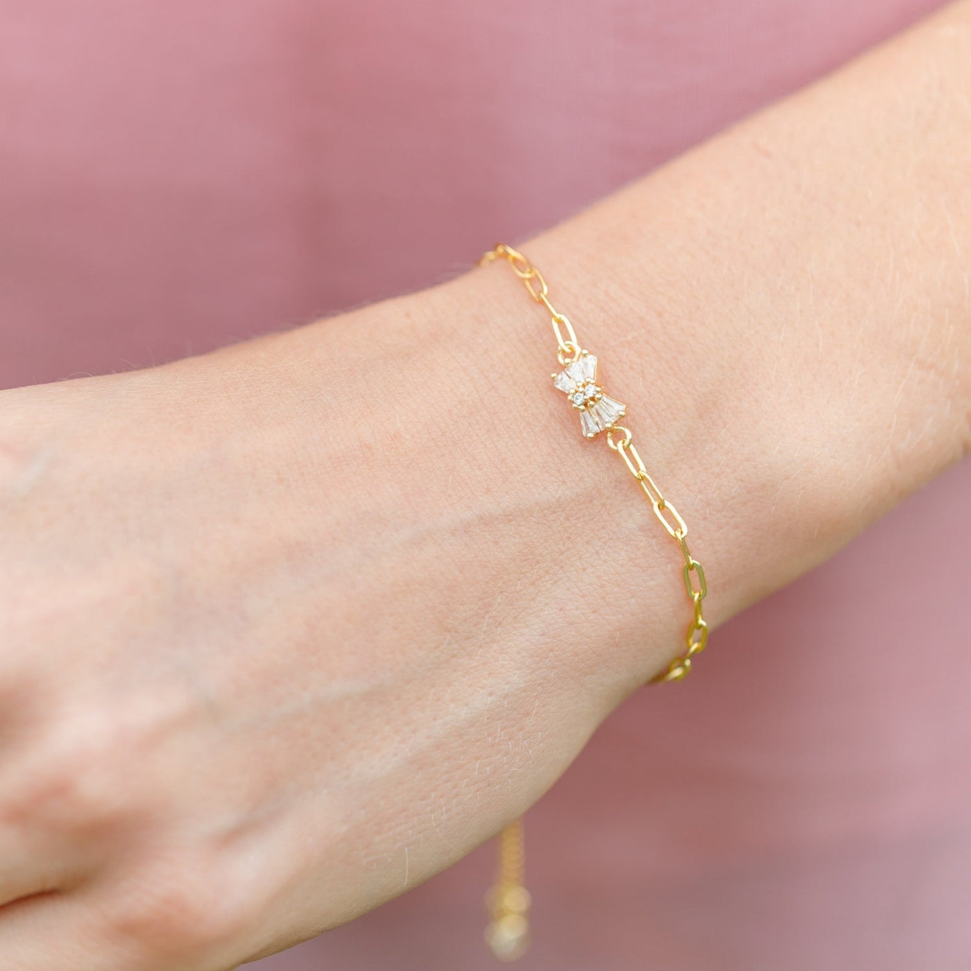Gold bracelet with a butterfly charm on a wrist against a pink background