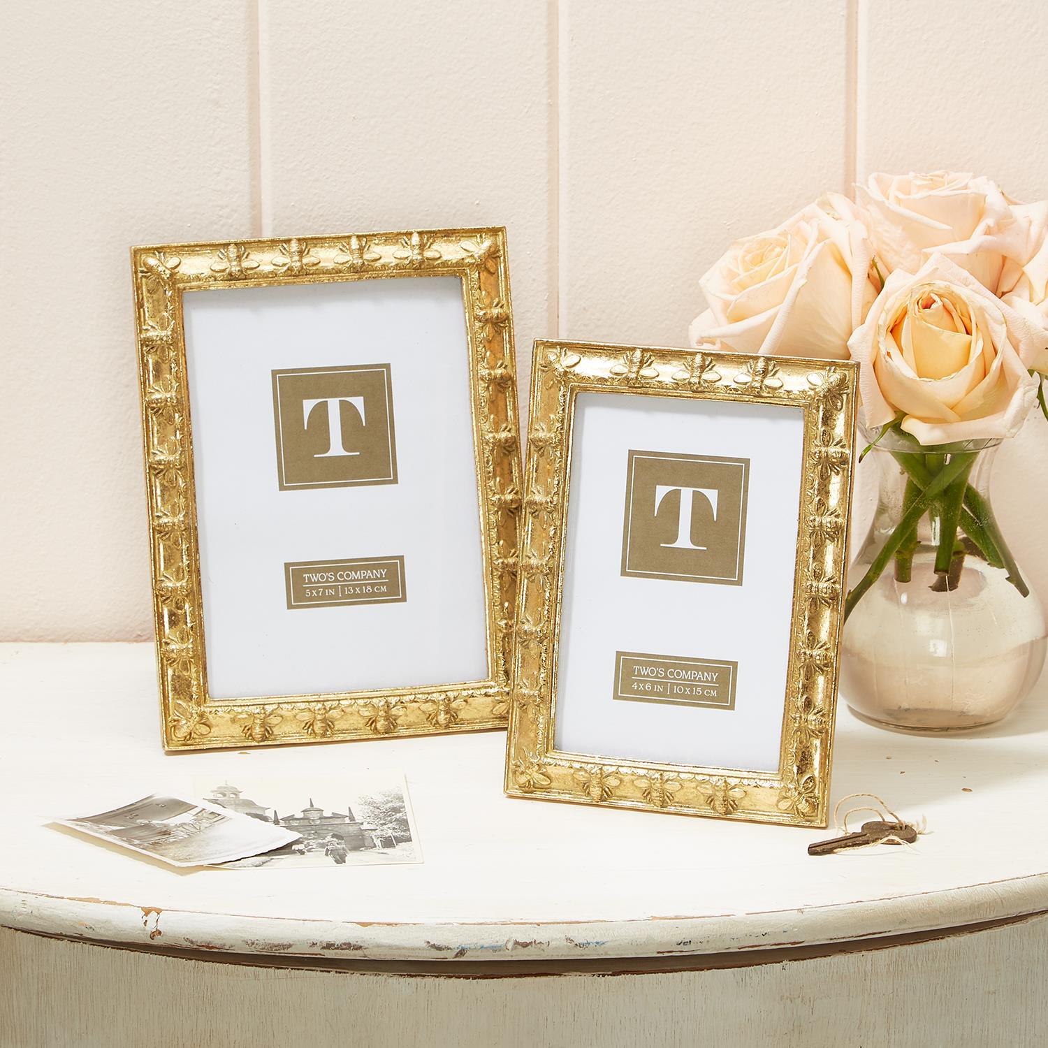 Two gold picture frames with a 'T' logo on a white surface with flowers in the background.