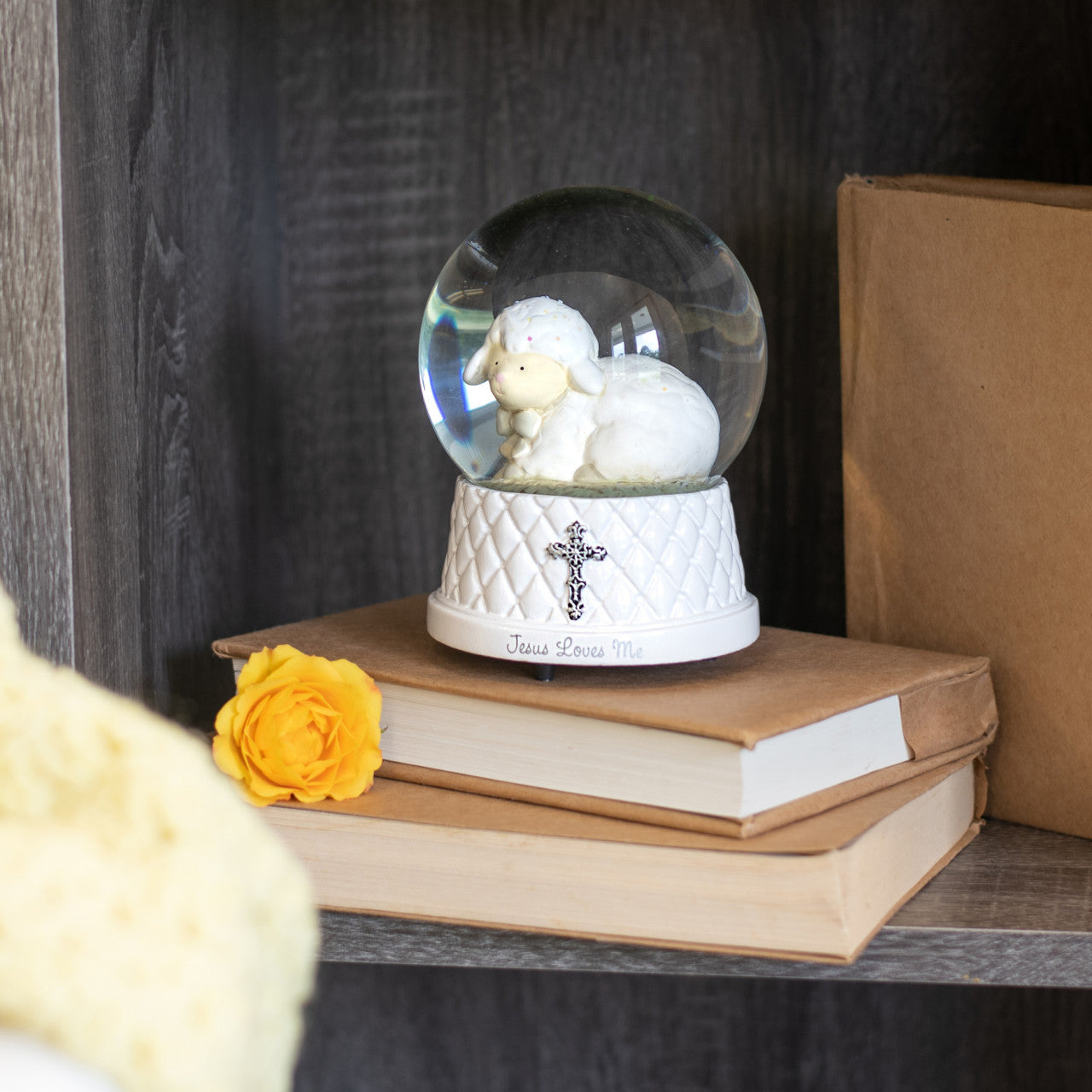 Decorative snow globe with sheep on top of books, featuring a cross and 'Jesus Loves Me' text.