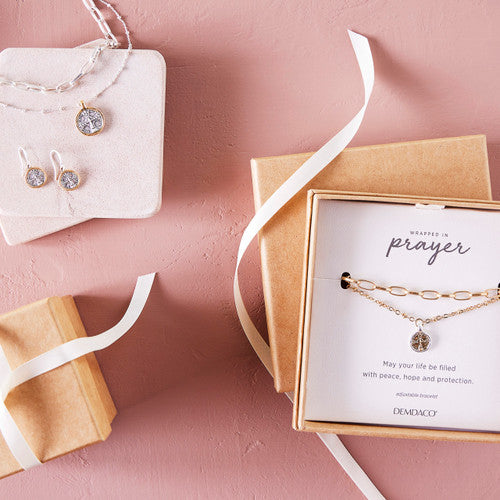 Gold necklace on a card with 'Prayer' text, placed on a pink surface with jewelry display.