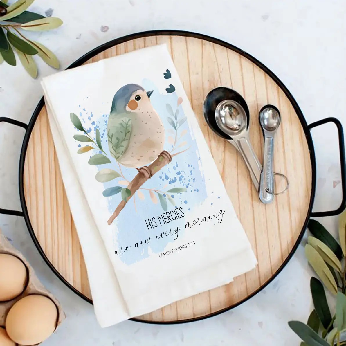 Tea towel with bird illustration and text on a wooden board with measuring spoons and eggs.