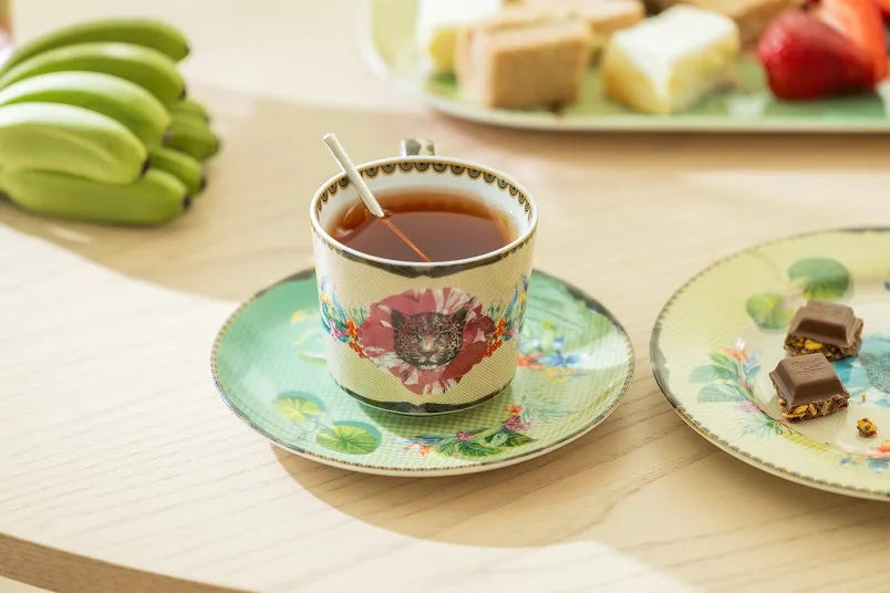 Teacup with floral design on a saucer, surrounded by snacks and fruits on a wooden table.