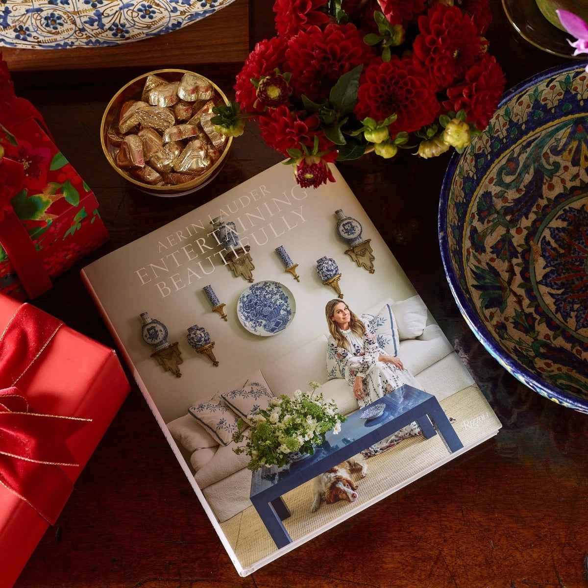 Book titled 'Decorating with Beautifully' on a table with decorative items including a bowl of cookies, flowers, and a colorful plate.