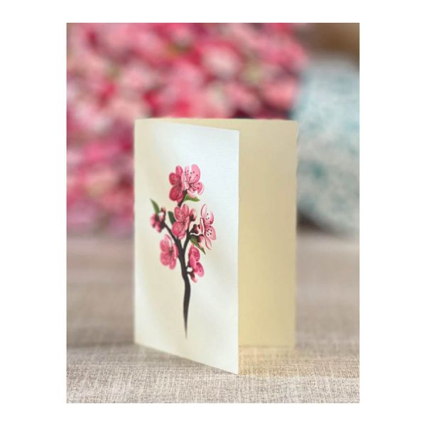 Candle with cherry blossom design on a white card, placed on a textured surface with blurred pink flowers in the background.