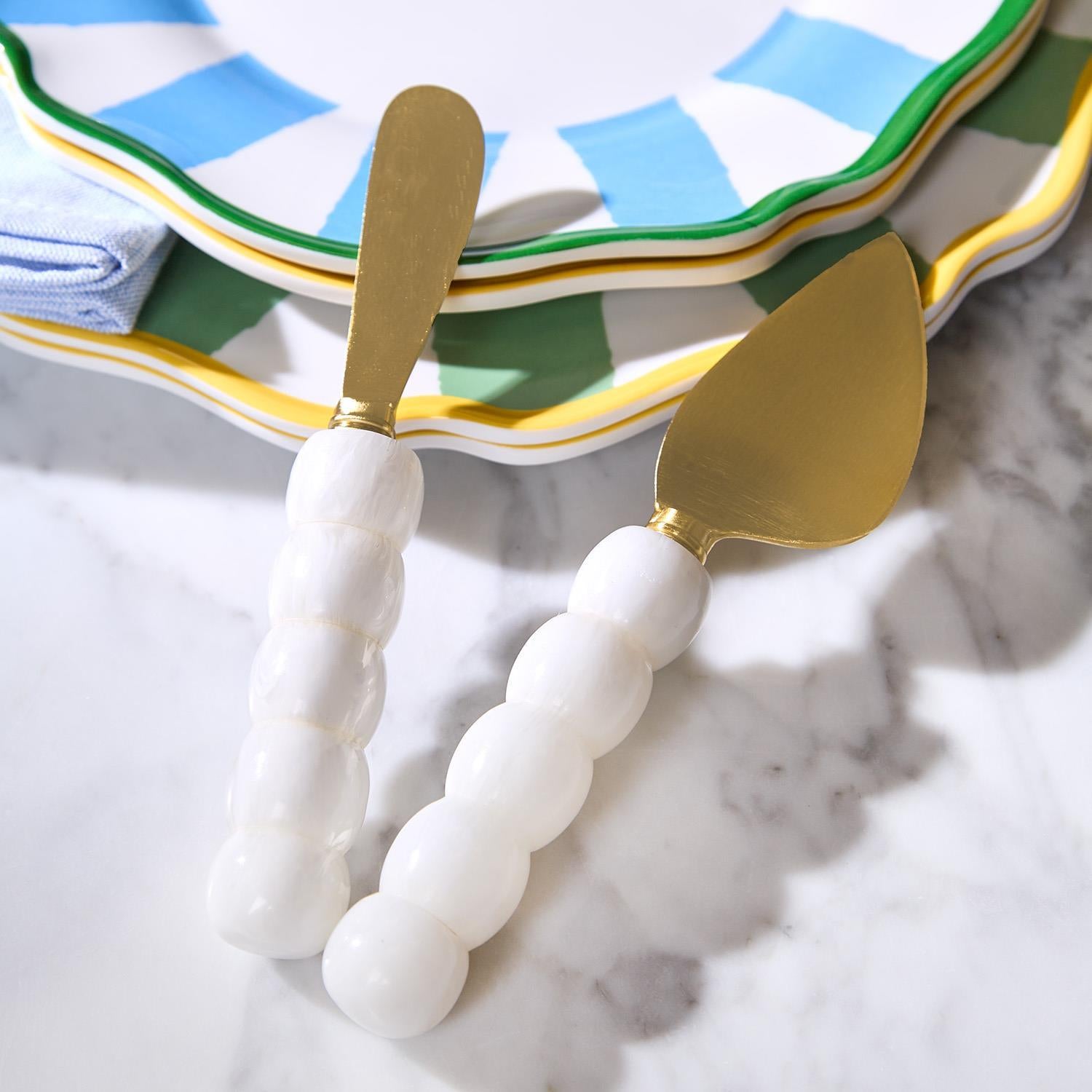 Two cheese knives with white chunky ball handles and gold blades on a marble surface.