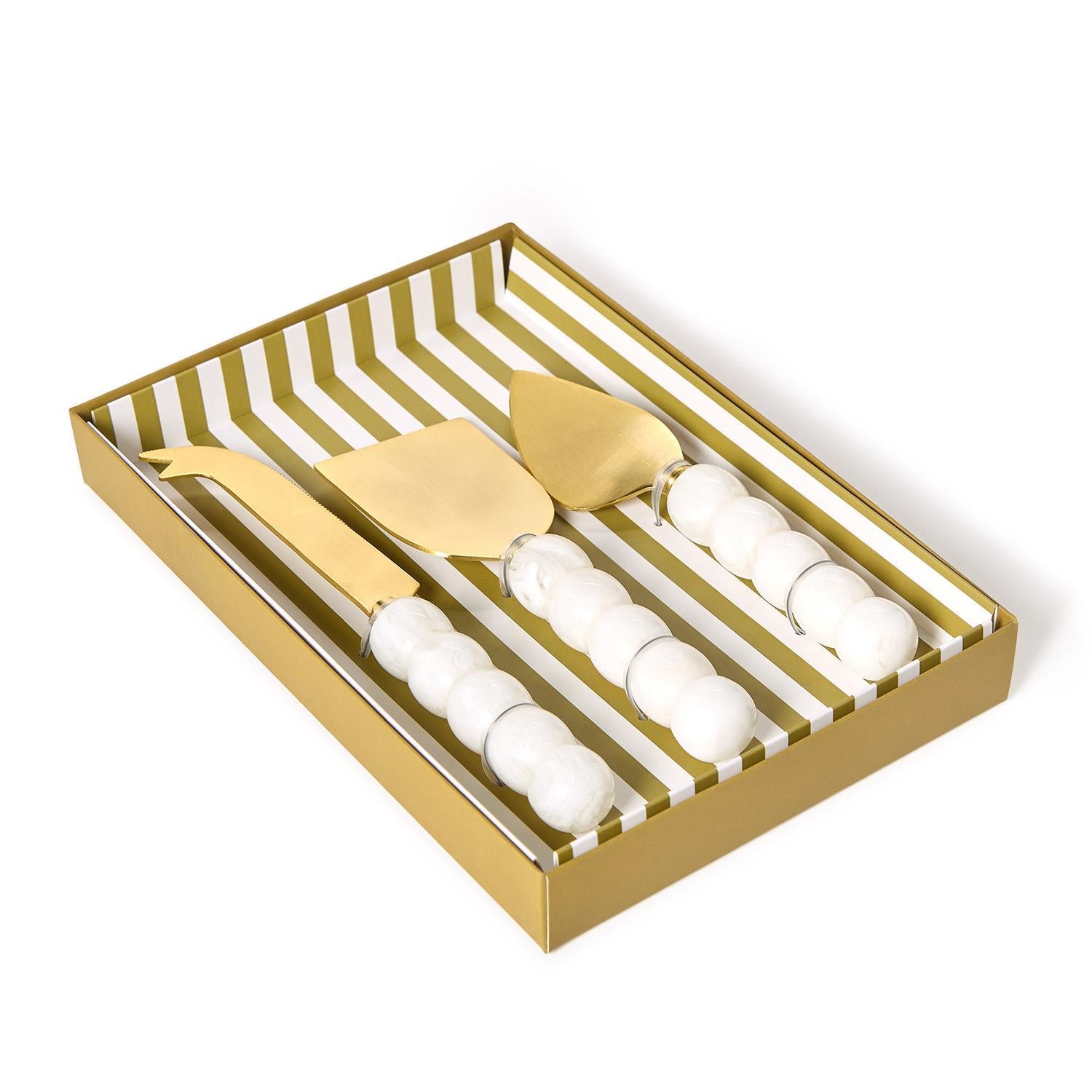 Gold cheese knife set with white chunky ball handles in a striped box on a white background