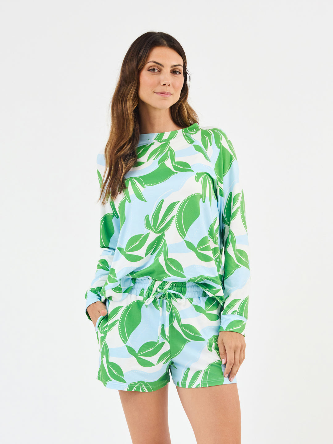 Woman wearing Mary Square In The Shade Pajamas, Nature inspired floral print shorts with long sleeve top. green, turquoise and white leaf-patterned on a white background