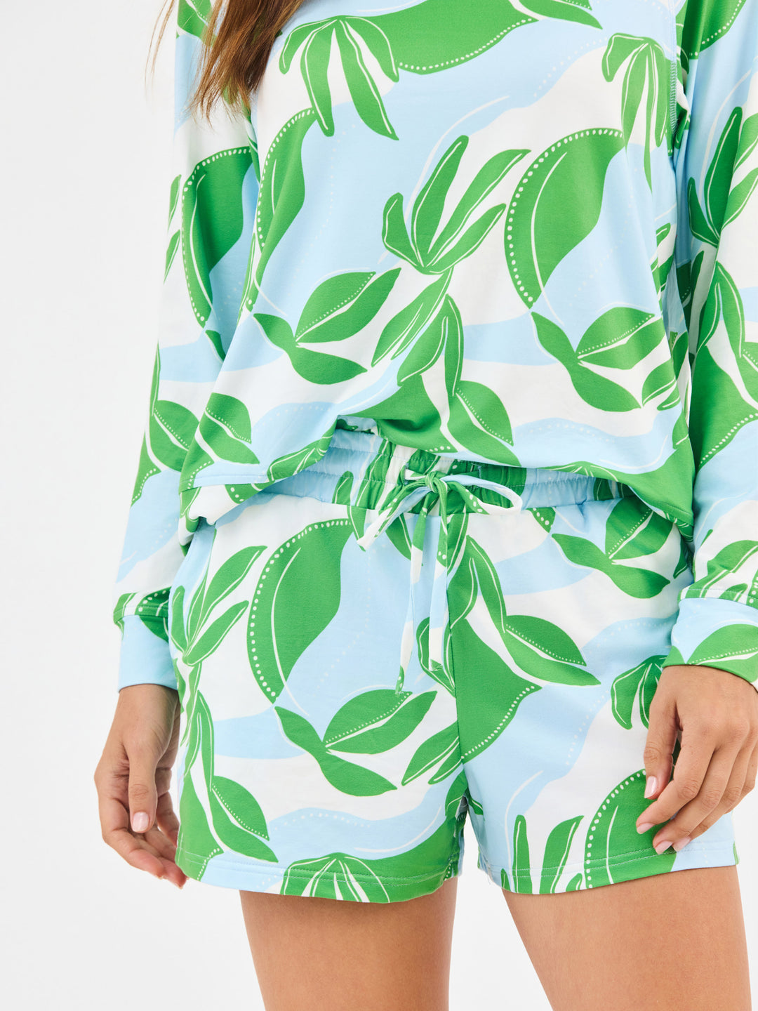Woman wearing Mary Square In The Shade Pajamas, Nature inspired floral print shorts with long sleeve top. green, turquoise and white leaf-patterned on a white background