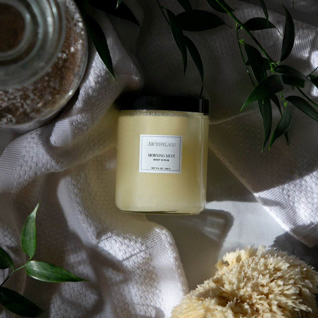 Jar of Archipelago Morning Mist body scrub on a textured surface with green leaves.