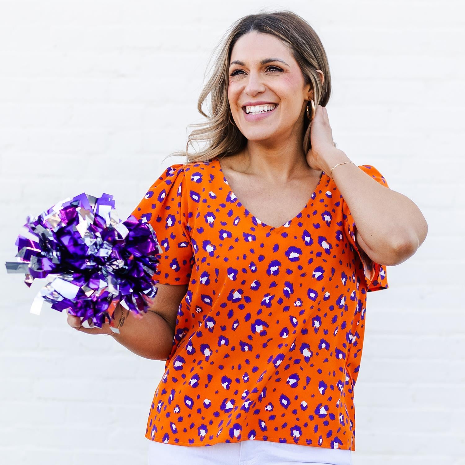 Woman wearing an orange blouse with purple leopard print holding purple and silver pom-poms against a white background