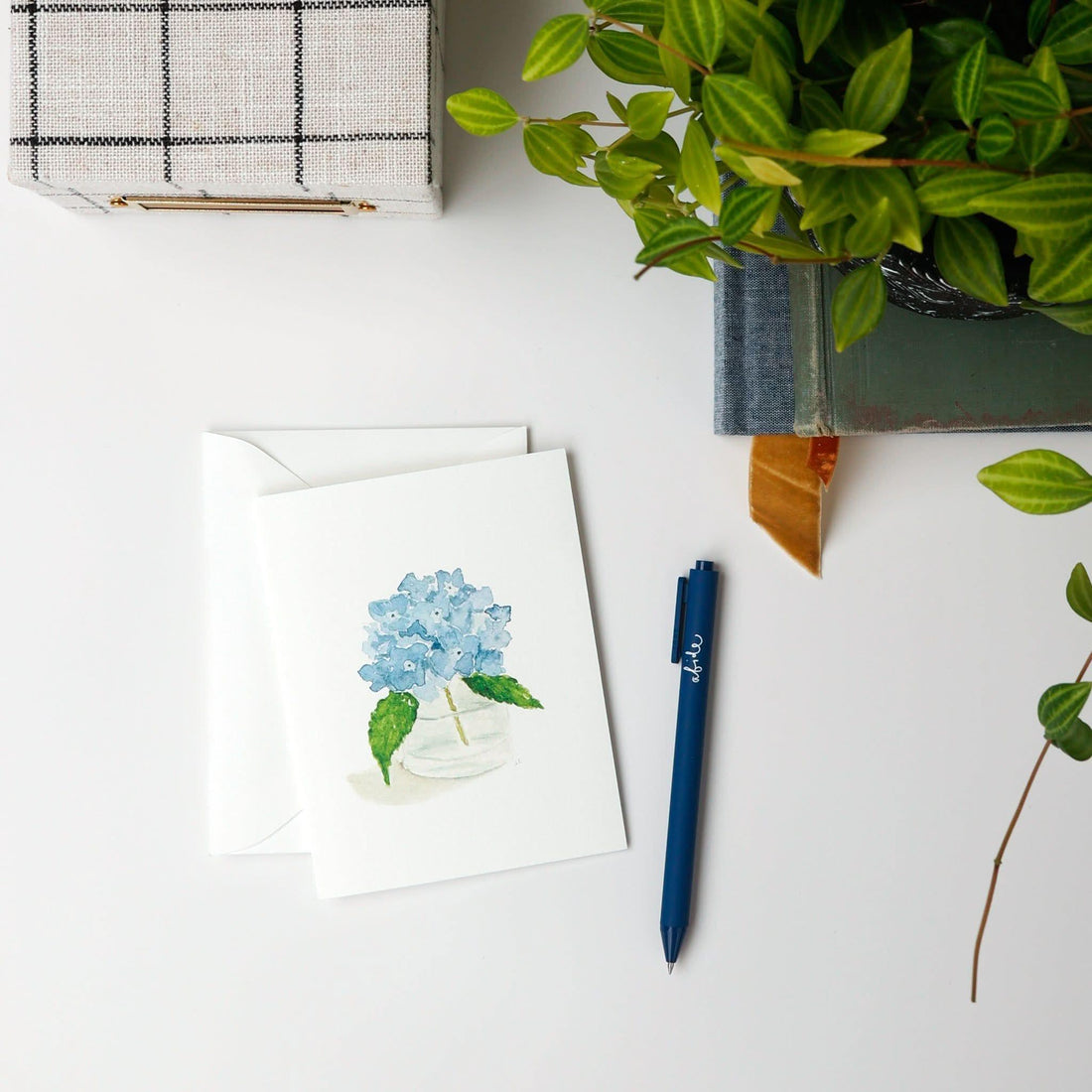 Greeting card with blue flower illustration, pen, and plant on a white surface