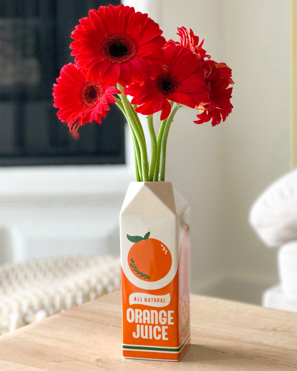 Red flowers in a vase designed like a carton of orange juice on a wooden surface