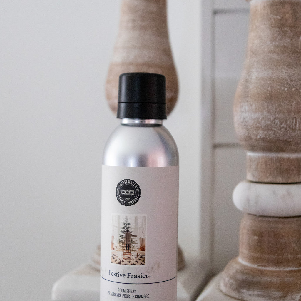 Seasonal - Room Spray - Festive Frasier scent.  Silver spray bottle with white label. Product is sitting on a table next to wooden candle sticks. 