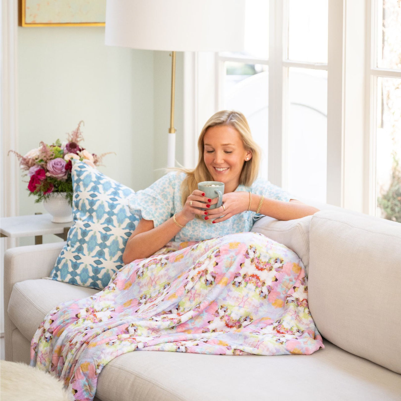 Woman sitting on a couch with a floral blanket, holding a mug in a bright living room.