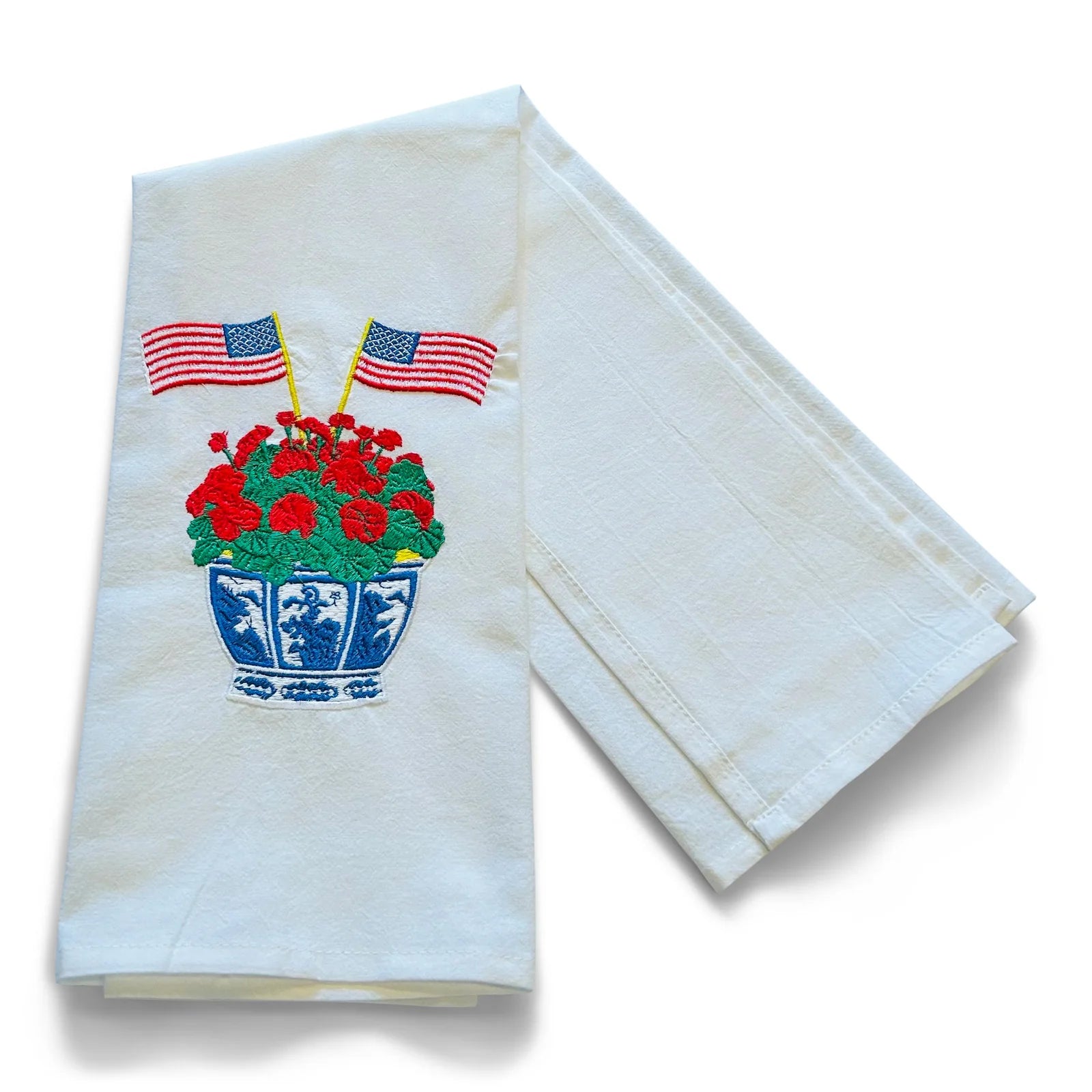 White towel with embroidered design of a flower pot with red flowers, blue pot, and two American flags on a white background.