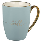 Light blue mug with gold handle and 'IT IS well' text on a white background