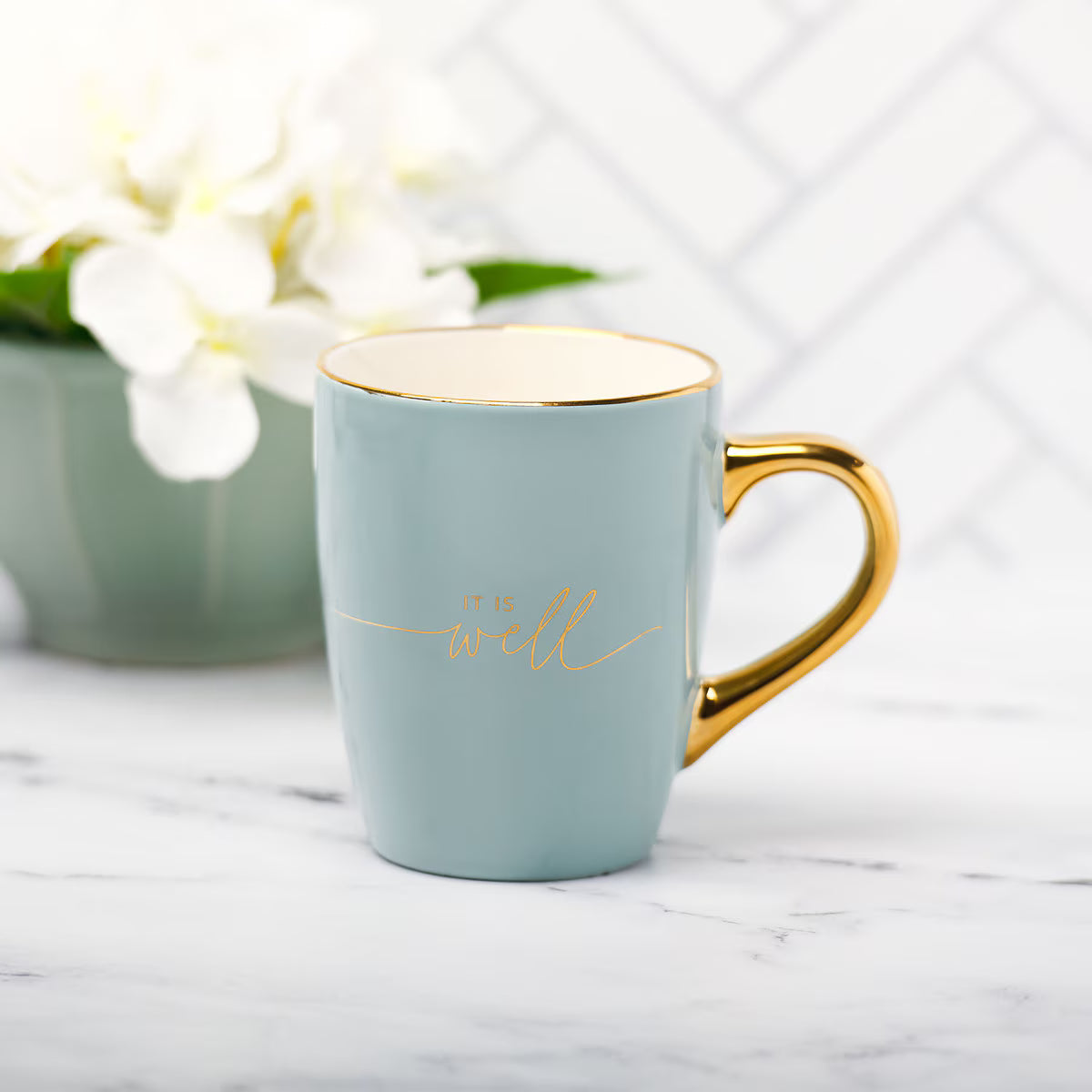 Light blue mug with gold handle and 'be well' text on a marble surface with flowers in the background