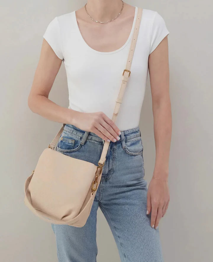Person wearing a white top and blue jeans holding a beige handbag against a plain background