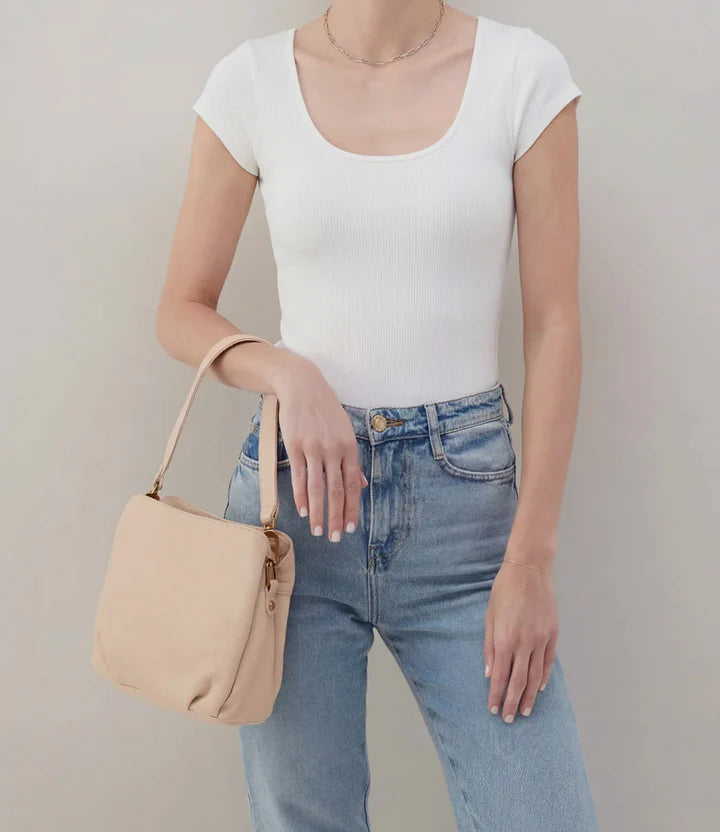 Person wearing a white t-shirt and blue jeans, holding a beige handbag against a plain background