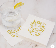 The Royal Standard Brand White 5-inch square cocktail napkins with gold tiger face print, styled on marble with an iced cocktail and lemon garnish"