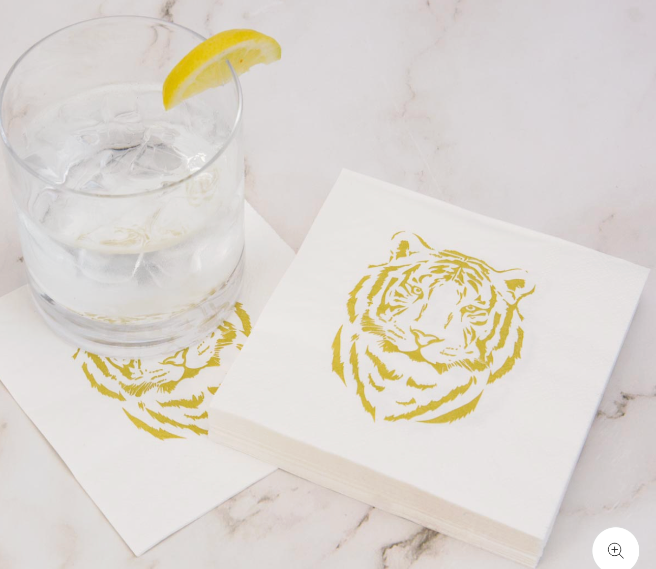 The Royal Standard Brand White 5-inch square cocktail napkins with gold tiger face print, styled on marble with an iced cocktail and lemon garnish"