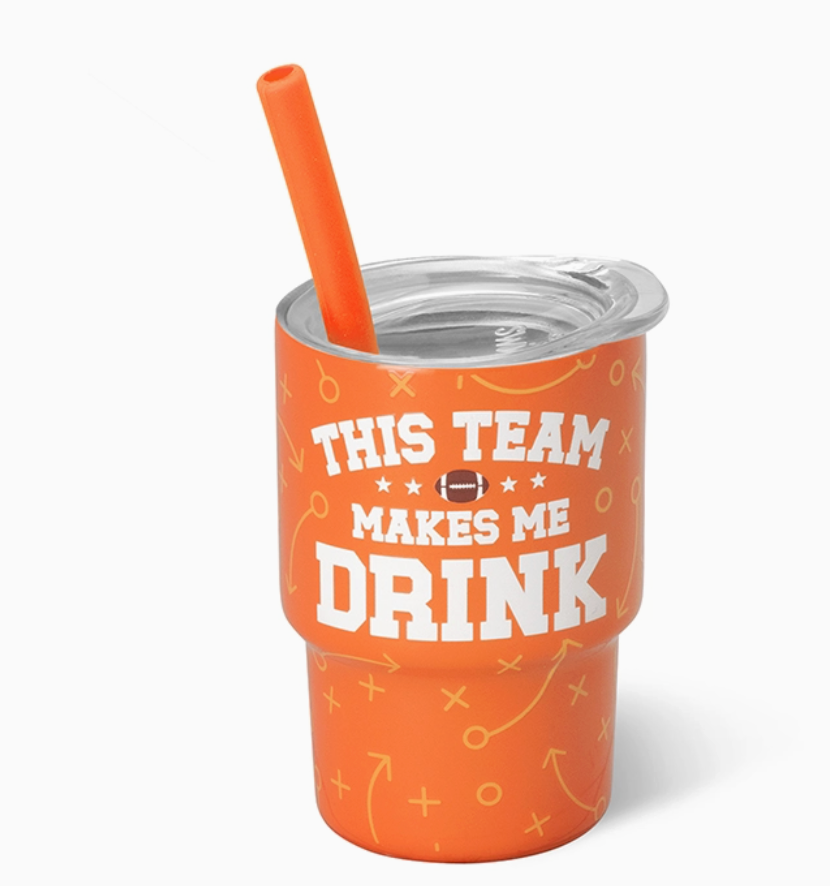 An orange insulated shot glass tumbler with a clear lid and matching orange straw, printed with the phrase 'THIS TEAM MAKES ME DRINK' in white text and small football-themed graphics, on a light gray background.
