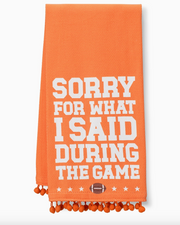 An orange woven kitchen towel with white text reading 'SORRY FOR WHAT I SAID DURING THE GAME' and orange pom-pom fringe along the bottom edge.