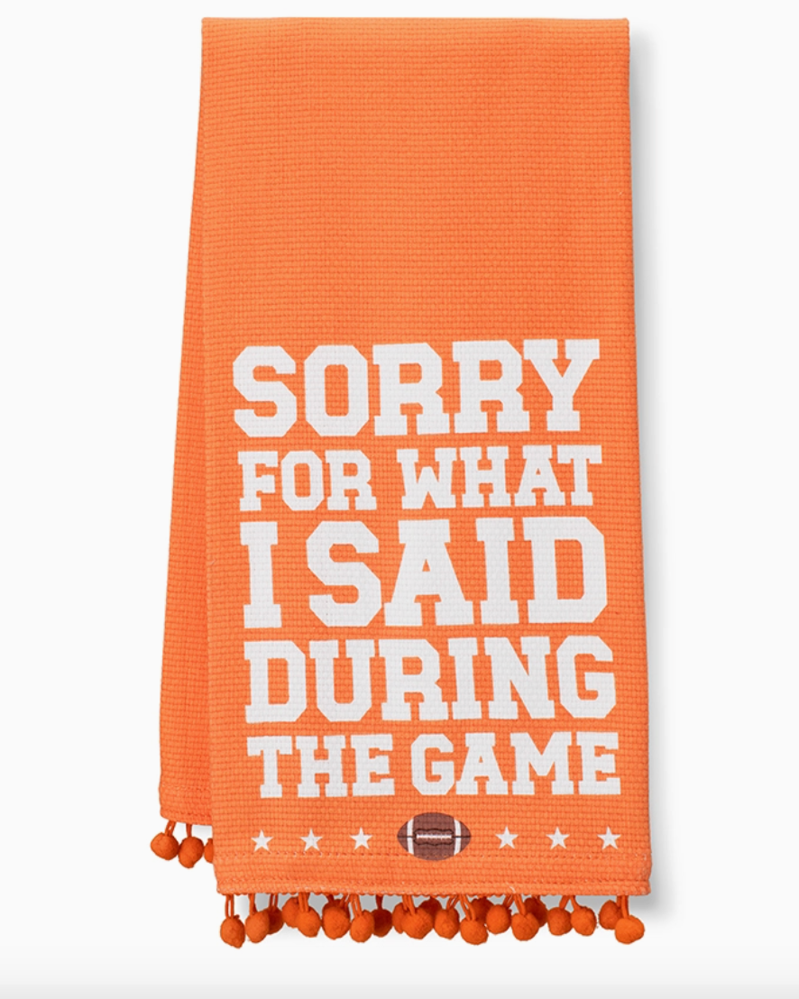 An orange woven kitchen towel with white text reading 'SORRY FOR WHAT I SAID DURING THE GAME' and orange pom-pom fringe along the bottom edge.