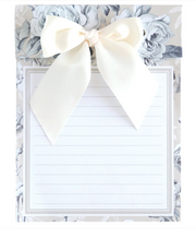 A lined notepad with a large cream satin bow on a floral-patterned backing, photographed against a white background.