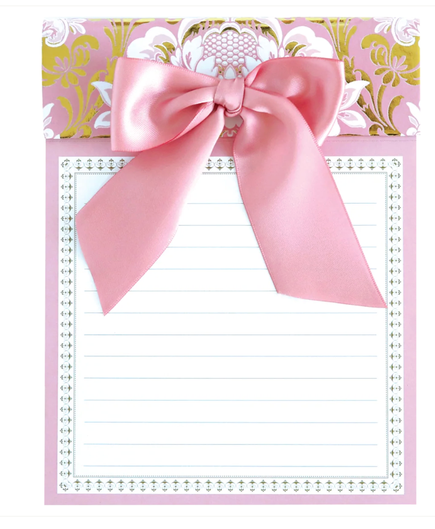 An Anna Grifin brand pink lined notepad with a large satin pink bow across the top, set against a decorative pink, white, and gold patterned top