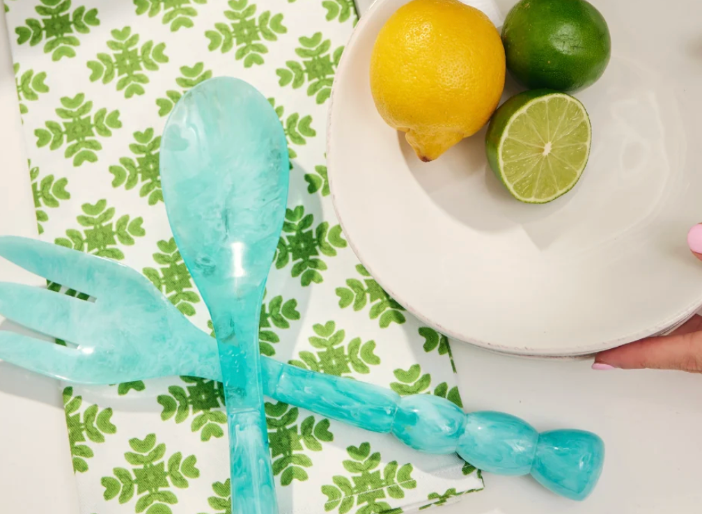 A Laura Park turquoise blue marbled resin serving utensil set featuring a spoon and a three-tine serving fork, both with decorative beaded handles, shown on a green print cloth with white bowl holding lemon and lime