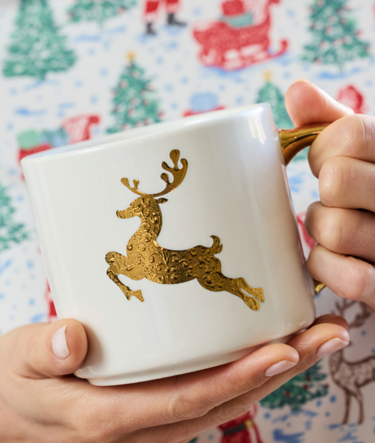 A white ceramic mug with a gold glittery reindeer design, held in two hands against a festive Christmas-themed background.
