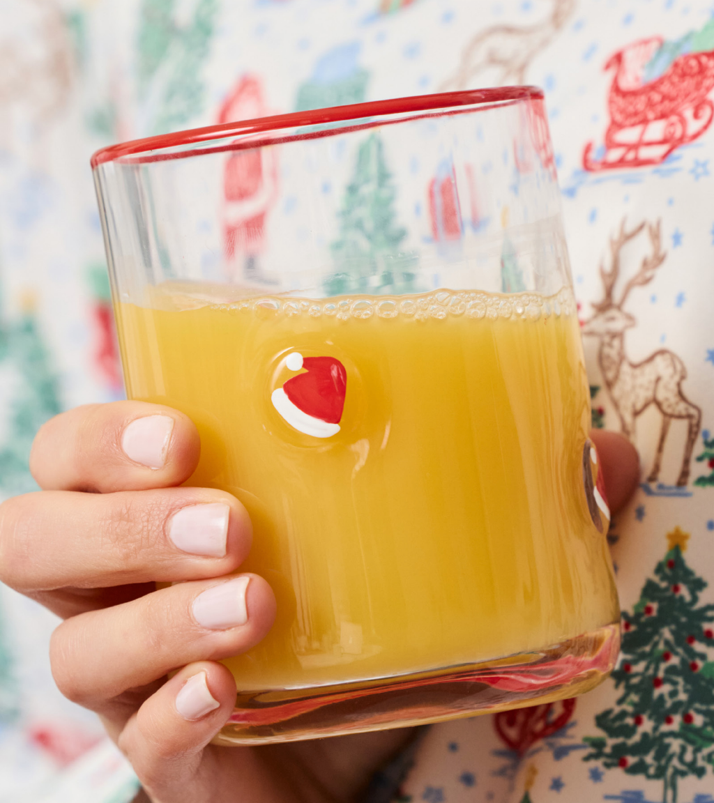 A hand holding a clear glass tumbler with a red rim and a small Santa hat detail, filled with orange juice, against a festive Christmas print background.