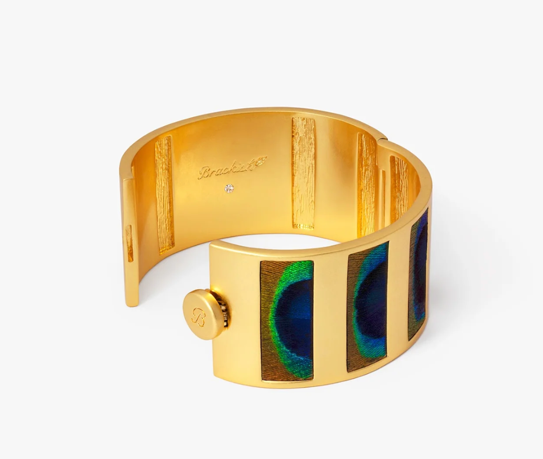 Brackish Natalie peacock feather cuff shown open, revealing its gold interior, hinge, and round button closure”
