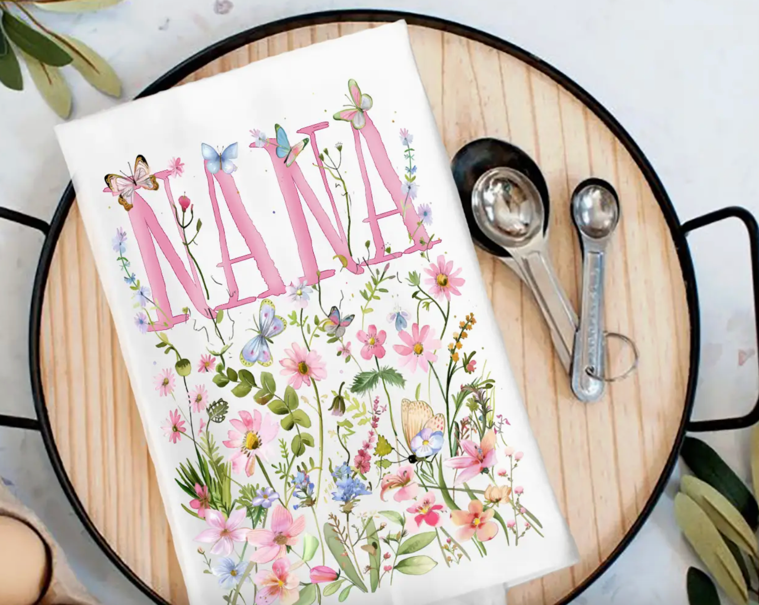 Floral-themed kitchen towel with 'Nana' printed on it, placed on a wooden cutting board with measuring spoons.