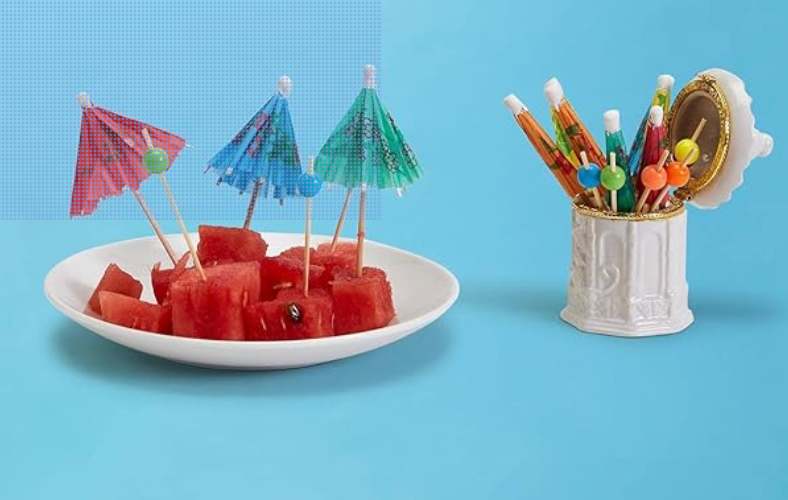 Watermelon slices with cocktail umbrellas picks on a plate and  more closed umbrella picks with  some with just a bead in a white ceramic pagoda jar against a blue background