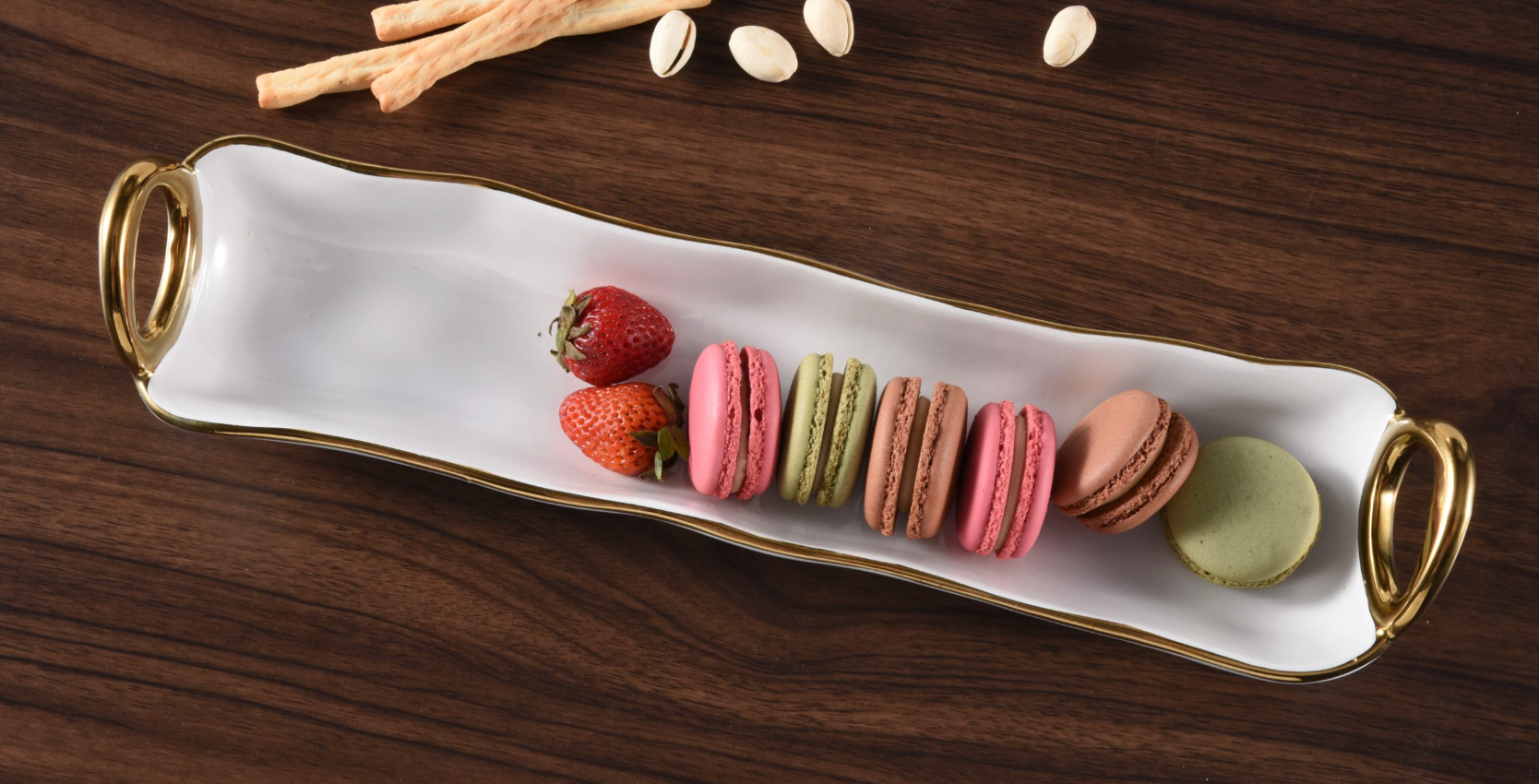Rectangular white tray with gold handles holding macarons and strawberries on a wooden surface.
