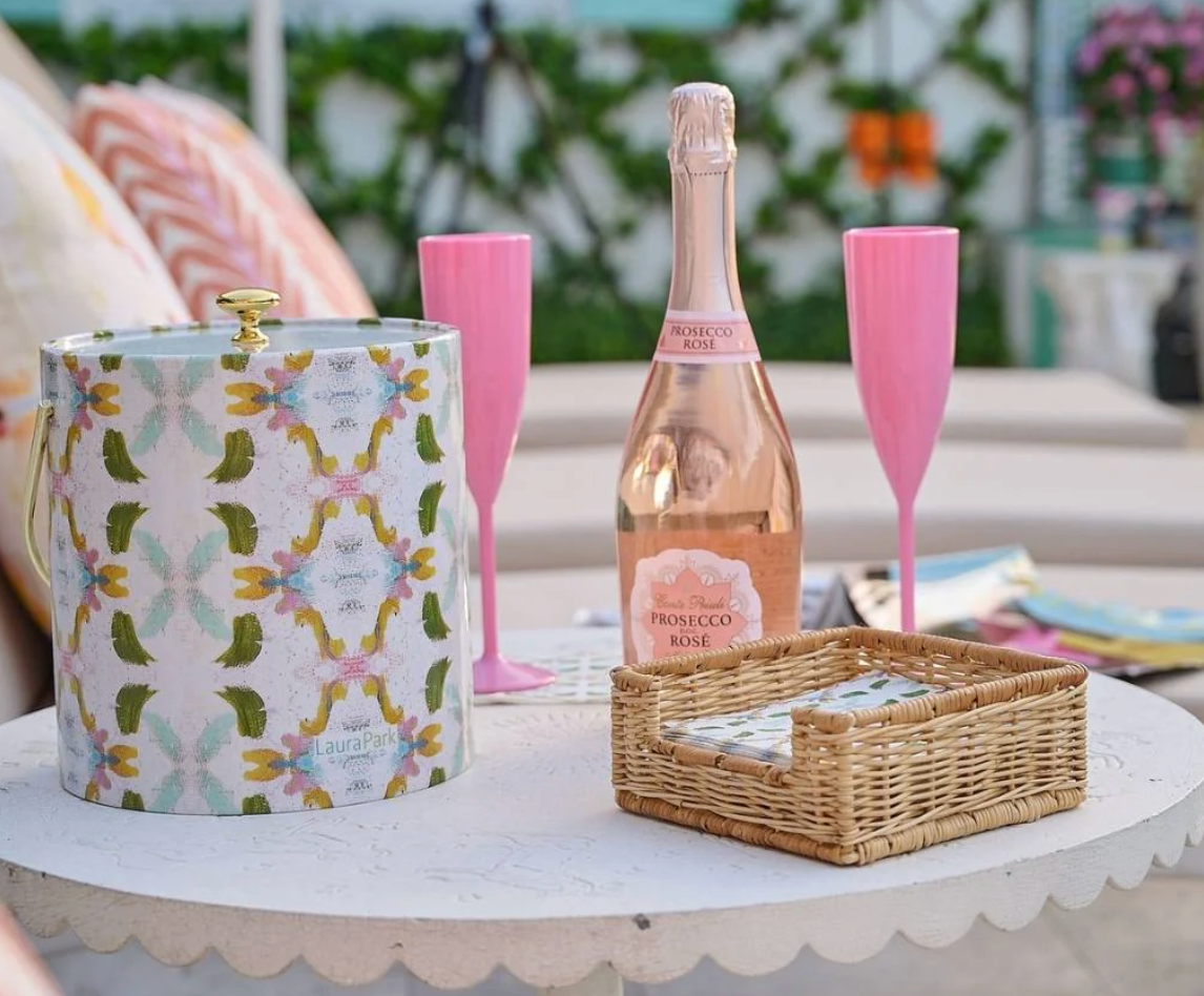 LAURA PARK DOGWOOD ICE bucket on table  and Bottle of Prosecco Rosé with two pink flutes and a decorative box on a table outdoors.