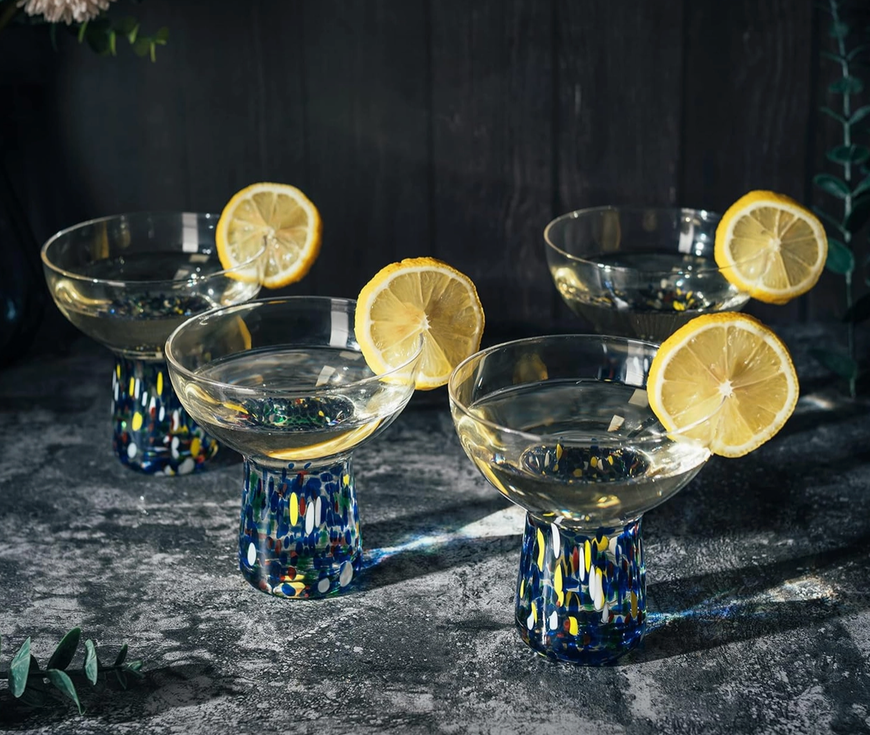 Set of 4 stemmed cocktail glasses with colorful confetti stems andbowl top with drink and lemon slices on a dark surface