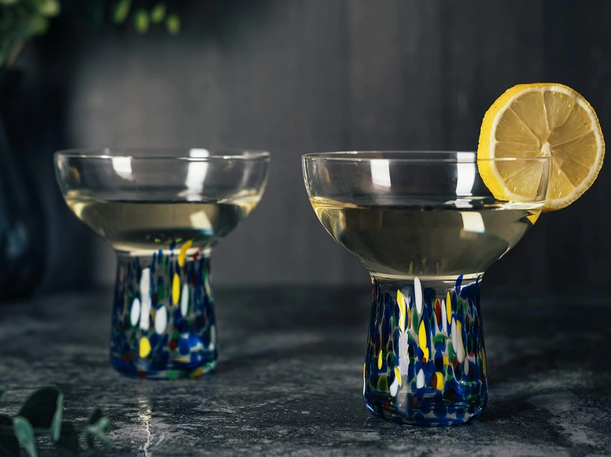 Two cocktail glasses with colorful bases and lemon slices on a dark surface.