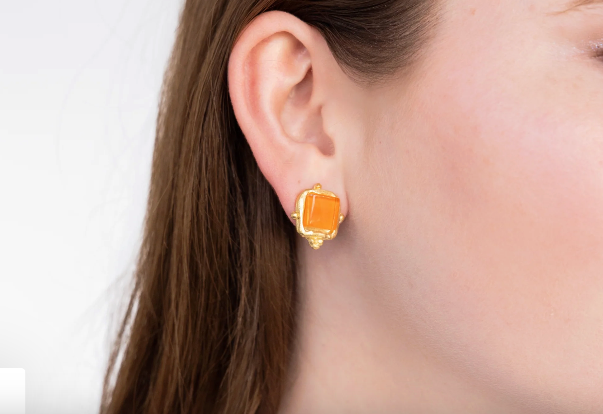 Close-up of a person wearing Susan Shaw  Charlotte style stud gold earrings with orange stones on a white background