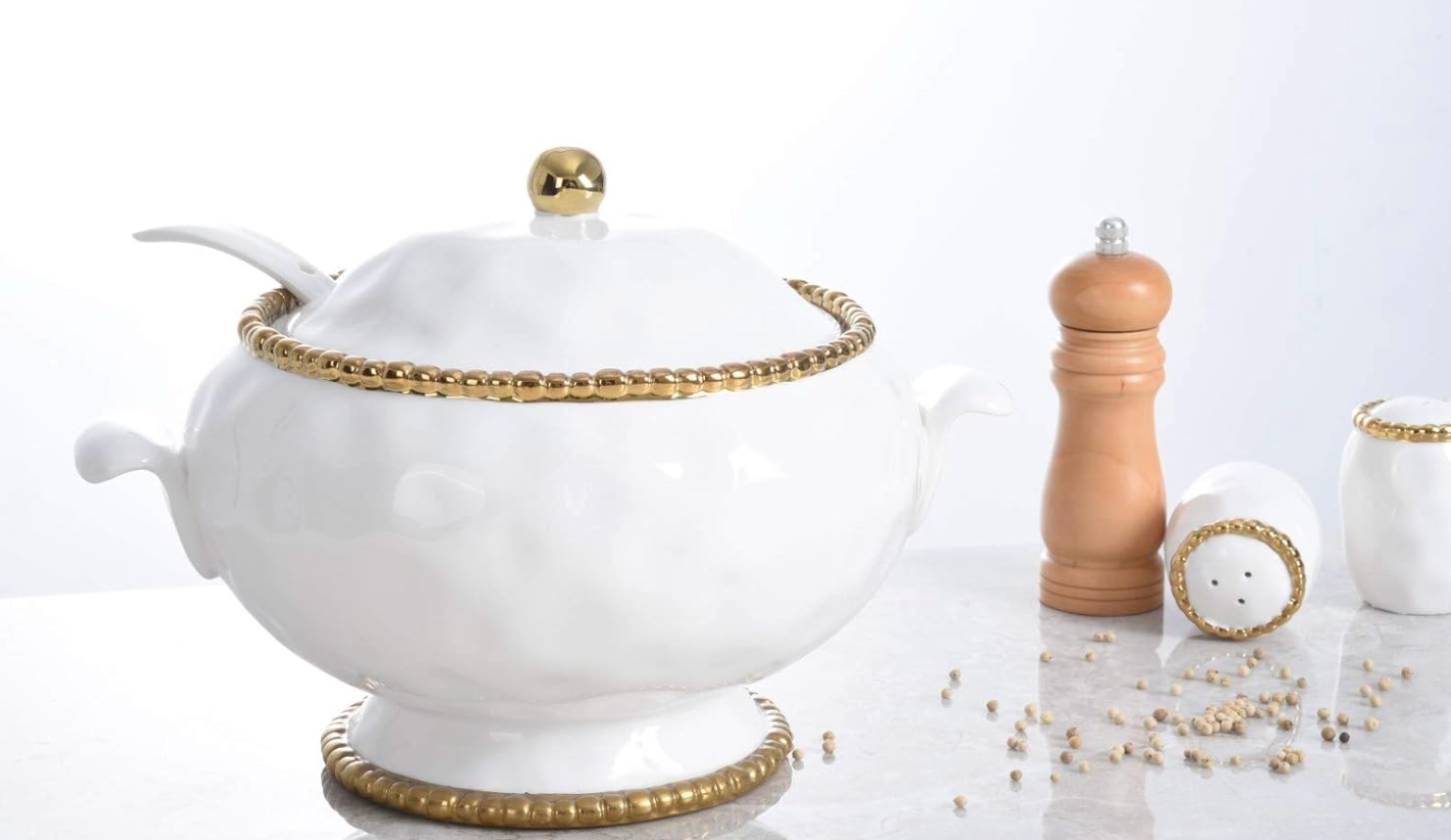 White ceramic and titanium Pampa Bay tureen, lid and spoon with gold accents on a light gray surface with salt and pepper in the background.