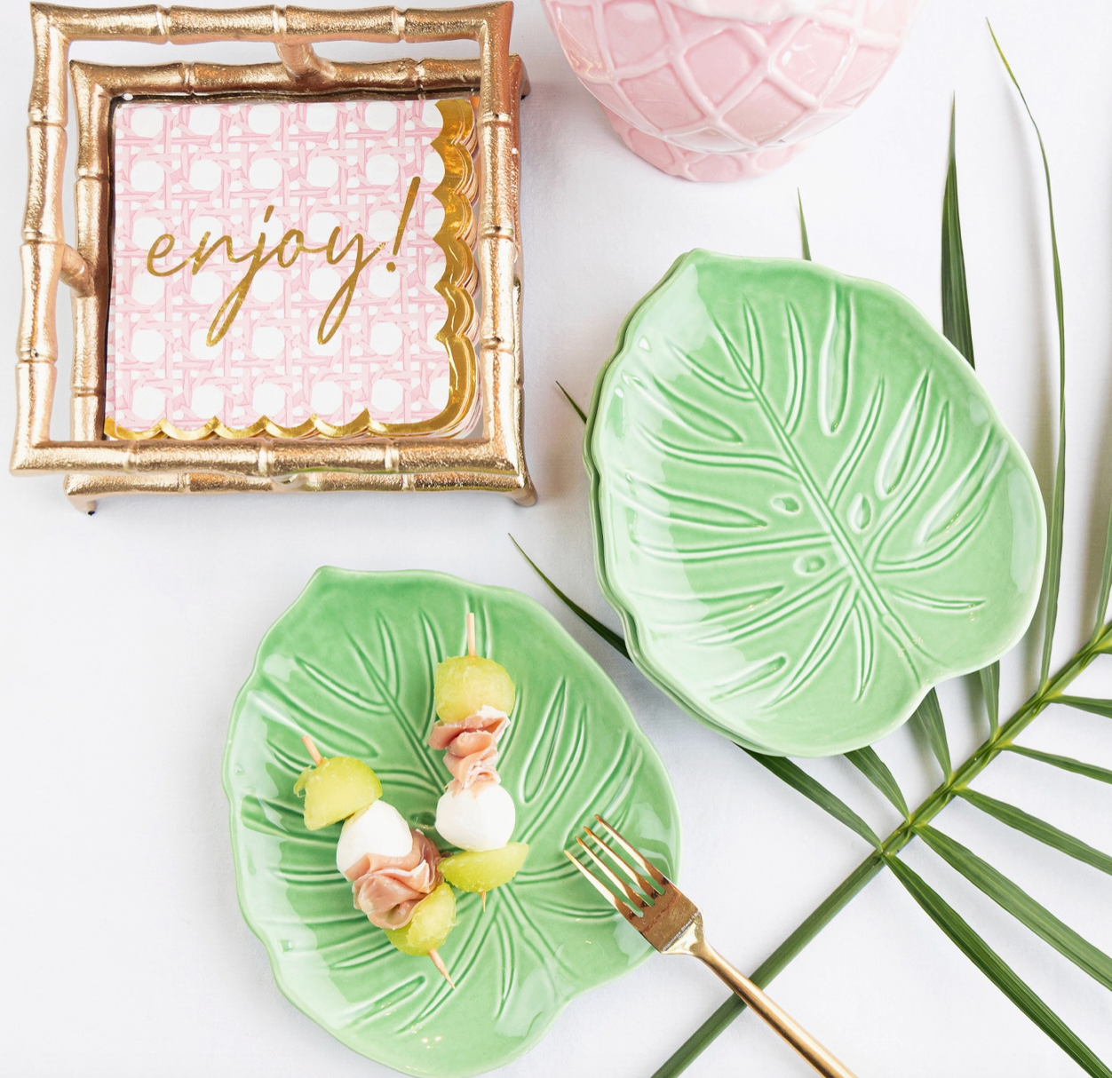 8 Oak Lane  gold bamboo cocktail napkin holder with  napkins that say enjoy! along side Green leaf-shaped plates with a small dessert and a gold fork.