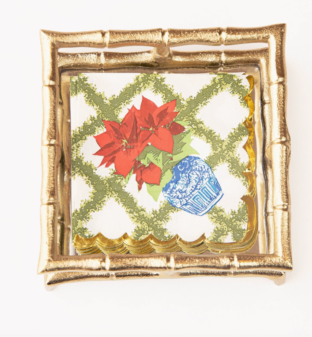 Decorative Christmas 8 Oak Lane Cocktail napkins with red poinsettias , a  green trellis. pattern and a blue vase on a white background with gold leaf effect scallop edge sitting in a gold bamboo napkin holder